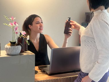 Spa ya da güzellik salonunda müşteriyle konuşan genç bir resepsiyonist. Masada losyon şişeleri var..