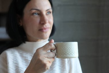 Orta yaşlı kadın pencerenin kenarında duruyor, kahve ya da çay içiyor, sessizliğin tadını çıkarıyor..