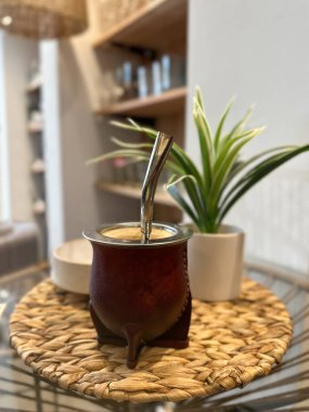 Sıcak, nötr tonlu bir iç mekanda, bir masaya samanla eş bardağının yakın plan çekimi. Yumuşak, bej, ahşap, pastel elementler ve ev bitkileriyle çevrili.