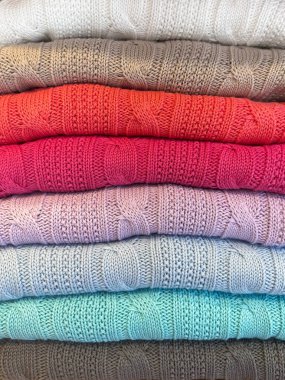 Düzgünce katlanmış kıyafetlerin yakın çekimi, sadece belli aralıklarla görünen renkleri gösteriyor. Eşgüdümlü tonlarda yumuşak, dokulu bir eğim oluşturan süveter ya da örgü gibi..