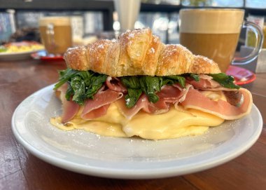 Kızarmış peynirli kruvasan sandviçi, jambon ya da domuz pastırması ve taze roka, bir sokak kafeteryasındaki ahşap masada servis edilir. Yanında kapuçino ya da sütlü kahve var..