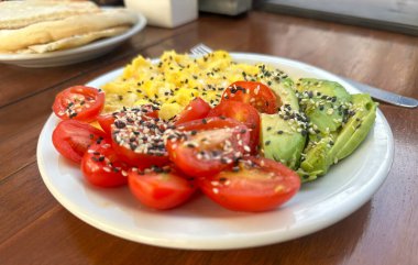 Kafe veya restoran ortamında sunulan sağlıklı bir kahvaltıya yakın çekim: omlet, dilimlenmiş domates ve tahtadan bir masada, tabakta tohum kaplı avokado..