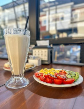 Kafe ya da restoran ortamında sunulan sağlıklı bir kahvaltıya yakın plan: omlet, dilimlenmiş domates ve tabakta tohum kaplı avokado, milkshake, hepsi ahşap bir masa üzerinde..