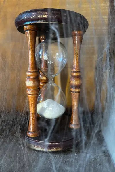Antik ahşap kum saati, alta yerleştirilmiş, örümcek ağlarıyla örtülmüş. Unutulmuş zaman ve çürüme için güçlü bir görsel metafor.,