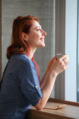 Kızıl saçlı kadın, 30 'lu yaşlarda, sıradan kıyafetlerle bir masada oturuyor, kahve içiyor ve pencereden dışarı bakıyor. Bir kafede ya da evde samimi, yansıtıcı bir an.