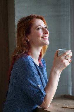 Kızıl saçlı kadın, 30 'lu yaşlarda, sıradan kıyafetlerle bir masada oturuyor, kahve içiyor ve pencereden dışarı bakıyor. Bir kafede ya da evde samimi, yansıtıcı bir an.