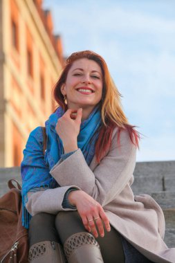 Kızıl saçlı kadın, 30 'lu yaşlarda, açık havada merdivenlerde oturuyor, paltosu ve atkısı ile neşeli ve şık. Sonbahar ya da ilkbahar doğal ışıkta portresi.