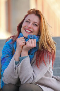 Kızıl saçlı kadın, 30 'lu yaşlarda, açık havada merdivenlerde oturuyor, paltosu ve atkısı ile neşeli ve şık. Sonbahar ya da ilkbahar doğal ışıkta portresi.