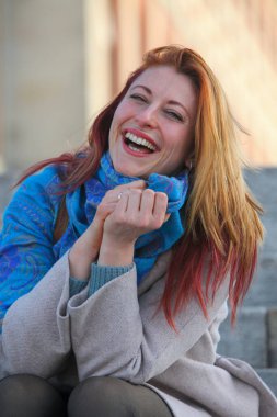 Kızıl saçlı kadın, 30 'lu yaşlarda, açık havada merdivenlerde oturuyor, paltosu ve atkısı ile neşeli ve şık. Sonbahar ya da ilkbahar doğal ışıkta portresi.