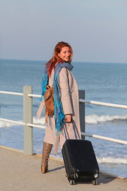 Kızıl saçlı kadın, 30 'larında, elinde bavuluyla iskelede duruyor. Palto ve eşarp içinde mutlu ve şık. Sonbahar ya da ilkbahar deniz havası.