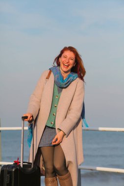 Kızıl saçlı kadın, 30 'larında, elinde bavuluyla iskelede duruyor. Palto ve eşarp içinde mutlu ve şık. Sonbahar ya da ilkbahar deniz havası.