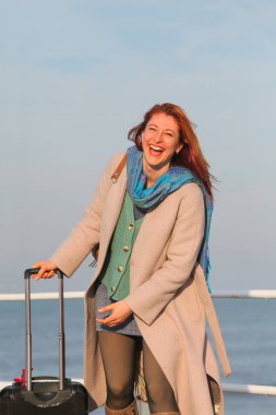 Kızıl saçlı kadın, 30 'larında, elinde bavuluyla iskelede duruyor. Palto ve eşarp içinde mutlu ve şık. Sonbahar ya da ilkbahar deniz havası.
