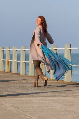 Kızıl saçlı kadın, 30 'lu yaşlarda, elinde atkıyla deniz kenarında yürüyor. Mutlu, şık, palto ve sonbahar ya da bahar kıyafetleri içinde. Neşeli ve kaygısız.