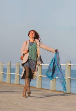 Kızıl saçlı kadın, 30 'lu yaşlarda, elinde atkıyla deniz kenarında yürüyor. Mutlu, şık, palto ve sonbahar ya da bahar kıyafetleri içinde. Neşeli ve kaygısız.