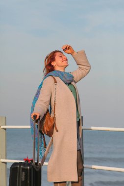 Kızıl saçlı kadın, 30 'larında, elinde bavuluyla iskelede duruyor. Palto ve eşarp içinde mutlu ve şık. Sonbahar ya da ilkbahar deniz havası.