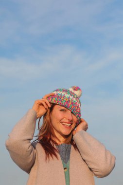 Oyuncu kızıl saçlı kadın, 30 'lu yaşlarda, deniz kenarında, renkli bir kıyafeti ve komik şapkası var. Gülerek ve aptalca davranarak. Parlak, neşeli bahar ya da sonbahar havası.