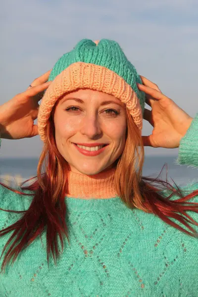 Oyuncu kızıl saçlı kadın, 30 'lu yaşlarda, deniz kenarında, renkli bir kıyafeti ve komik şapkası var. Gülerek ve aptalca davranarak. Parlak, neşeli bahar ya da sonbahar havası.