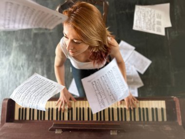 Kızıl saçlı kadın, 30 'larında, evde piyano çalıyor. Yüksek açılı bir manzara. Etrafında müzik notaları var ve yerde yatıyor. Sanatsal, duygusal ve anlamlı bir sahne..
