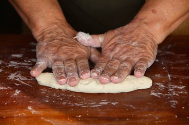 Yaşlı bir kadının ellerine yakın çekim, sıradan giyinmiş, basit bir ahşap masa üzerinde elle hamur yapıyor.
