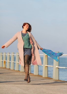 Kızıl saçlı kadın, 30 'lu yaşlarda, elinde atkıyla deniz kenarında yürüyor. Mutlu, şık, palto ve sonbahar ya da bahar kıyafetleri içinde. Neşeli ve kaygısız.