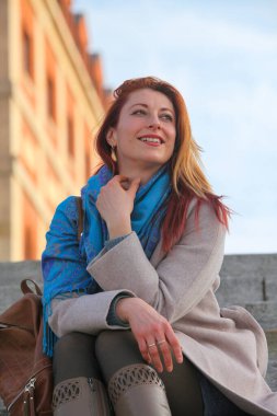  Kızıl saçlı kadın, 30 'lu yaşlarda, açık havada merdivenlerde oturuyor, paltosu ve atkısı ile neşeli ve şık. Sonbahar ya da ilkbahar doğal ışıkta portresi.