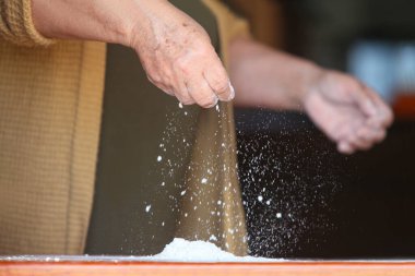 Yaşlı bir kadının ellerine yakın çekim, sıradan giyinmiş, basit bir ahşap masa üzerinde elle hamur yapıyor.