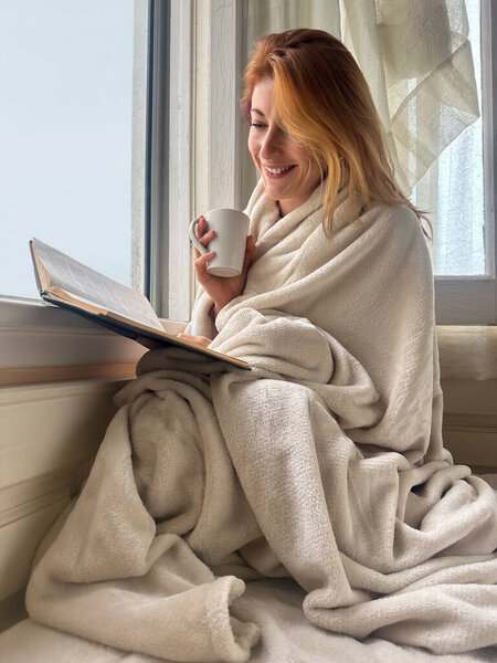Ginger-haired woman, 30s, sitting on bed by the window covered in blanket, reading a book and drinking coffee. Calm, peaceful moment in soft natural light.