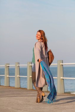 Kızıl saçlı, mutlu, 30 'lu yaşlarda, elinde fular, deniz kenarında yürüyen genç bir kadın. Mutlu, şık, palto ve sonbahar ya da bahar kıyafetleri içinde. Neşeli ve kaygısız.