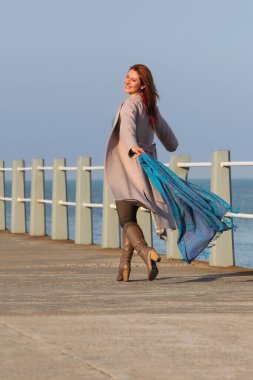 Kızıl saçlı, mutlu, 30 'lu yaşlarda, elinde atkıyla deniz kenarında yürüyen genç bir kadın. Mutlu, şık, palto ve sonbahar ya da bahar kıyafetleri içinde. Neşeli ve kaygısız.