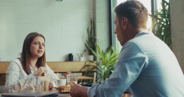 İki iş arkadaşı, bir erkek ve bir kadın, Chia ve salatayla sağlıklı bir öğle yemeği yerken rahat bir dostça sohbet ediyorlar. Ağır çekim, iş kafeteryasındaki rahat ve güneşli atmosferi yakalar..