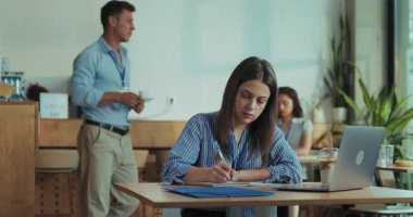 Kadın çalışan öğle yemeği molasında laptopunu, defterini ve kalemini kullanarak yavaş çekimde yavaş çekimde atış noktalarını hazırlamaya odaklanırken, iş arkadaşları arka planda hafif sohbetin keyfini çıkarıyorlar..