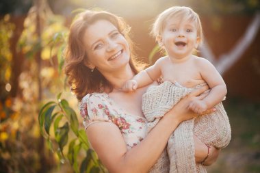 Genç bir kadın küçük oğlunu bahçede kollarında tutuyor. 