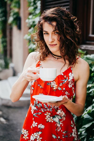 stock image Young beautiful woman in a red dress with a cup on  European steet