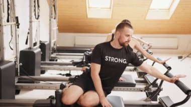 Yakışıklı sakallı erkek eğitmen bayan müşterilerini pilates reformist stüdyosunda sırayla otururken gösteriyor. Rehabilitasyon, sağlıklı yaşam tarzı. Büyük bir spor salonunda eğitmen..