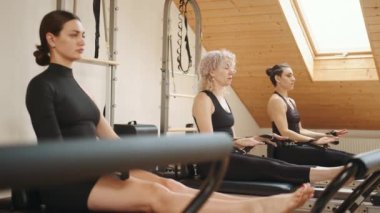 Bir grup aktif kadın, modern pilates stüdyosunda üst üste ıslah yatağında çalışırken tahta egzersizi yapıyor.