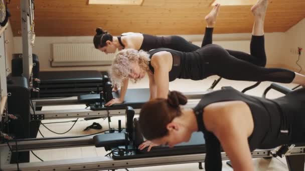 在现代普拉提工作室 妇女在更新改造床上锻炼时做木板运动 在现代普拉提演播室里 一组积极的妇女在改革床上连续锻炼时做着木板运动 — 图库视频影像