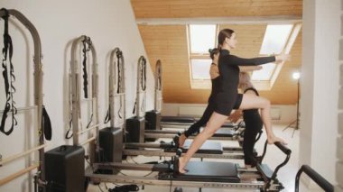 Spor merkezi, pilates stüdyosu. İki genç insan spor merkezinde kişisel antrenörleriyle antrenman yapıyor. Pilates stüdyosunun içinde. Yaşlı kadın eğitmen genç bayana yardım ediyor..