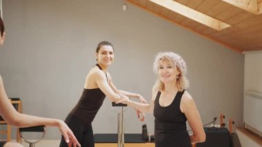 Sandalye üzerine pilates dersi, yaşlı sarışın eğitmenin stüdyoda iki genç kadına koçluk yapması. Kas iskeleti sisteminin düzeltilmesi, güzel bir vücut. Doğru duruş. Güneş ışığı efekti