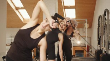 Siyah spor giyim giymiş esmer beyaz kadınlar, modern pilates stüdyosundaki parlak stüdyoda kişisel eğitmenleriyle reformist makineleri eğitimine direnç bantlarını çekiyorlar. Fitness konsepti. Rehabilitasyon.