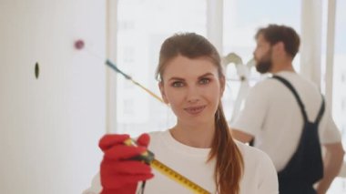 Young fun beautiful woman in casual clothes holding measure tape. Instruments, accessories for renovation apartment room. Repair home concept. In the background, a man paints a wall.