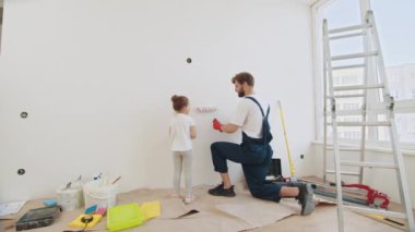 Smiling 4-year-old girl holds a paint roller in her hands. The child helps her father, who is standing behind her, near the wall, in the repair. Father and his daughter painting a wall in new home
