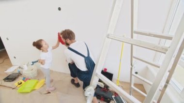 Smiling 4-year-old girl holds a paint roller in her hands. The child helps her father, who is standing behind her, near the wall, in the repair. Father and daughter give each other high fives