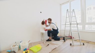 Father and daughter give each other high fives. Smiling 4-year-old girl holds a paint roller in her hands. The child helps her father, who is standing behind her, near the wall, in the repair