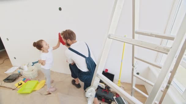 笑顔の4歳の少女は手にペイントローラーを持っています 子供は修理で 壁の近くに彼女の後ろに立っている彼女の父を助けます 父と娘はお互いに高い5を与える — ストック動画