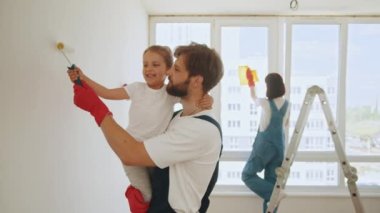 Family time together with their young daughter father, paint a wall with a roller. Joyful young Caucasian family painting room. Dad teaching little adorable daughter to paint wall using roller brush