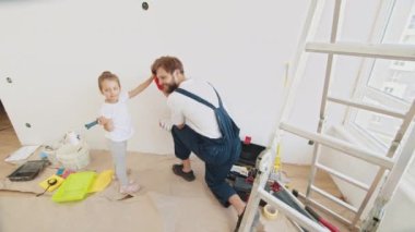 Smiling 4-year-old girl holds a paint roller in her hands. The child helps her father, who is standing behind her, near the wall, in the repair. Father and daughter give each other high fives