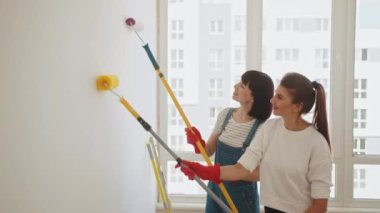 Two young women painting wall with paint roller while. Two attractive girlfriends paint the wall with a roller and a brush. LGBT family makes repairs inside the house. Female hands repair.