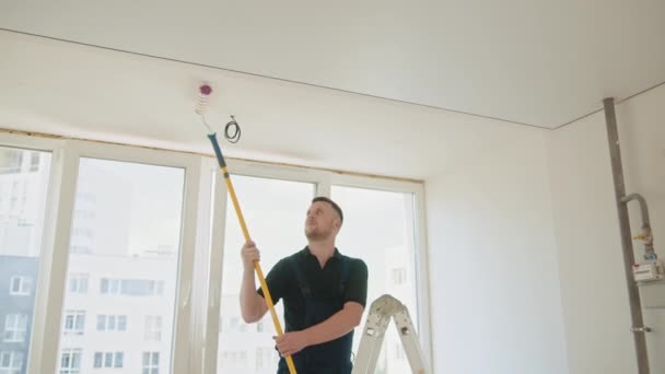その芸術家は人です 青の髭を生やした建設労働者がアパートの天井に絵を描いている ローラーを使ってる 男が新しいアパートの壁と天井を塗ります — ストック動画