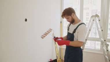 Duvarın rengini odadaki kumaşlardan seçen bir adam. Onarım, bina, teknoloji ve ev konsepti - erkeğe yakın çekim. Genç bir adam boya silindirini ve boş duvara karşı kumaş örneklerini tutuyor.