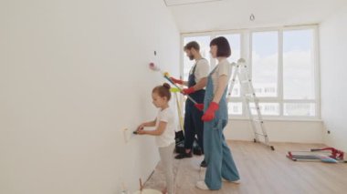 Genç Baba ve Anne, Çocuklarına Bir Duvar Nasıl Boyanır Gösteriyorlar. Boya rengi açık mavi. Evdeki oda tadilat için hazırlandı. Apartmanda tamirat var. Birlikte duvar boyuyoruz.